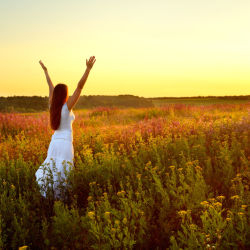 A woman standing in a field with her arms in the air looking at a sunset to illustrate a success concept
