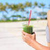 Woman on a beach holding a green smoothie getting into shape for summer