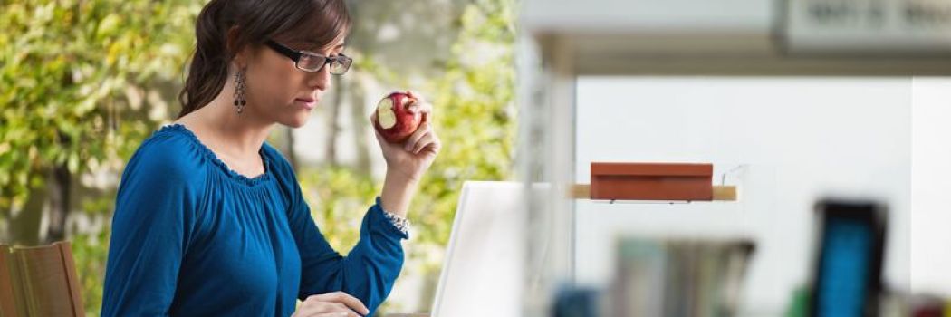 A woman sitting eating an apple while she works on a laptop